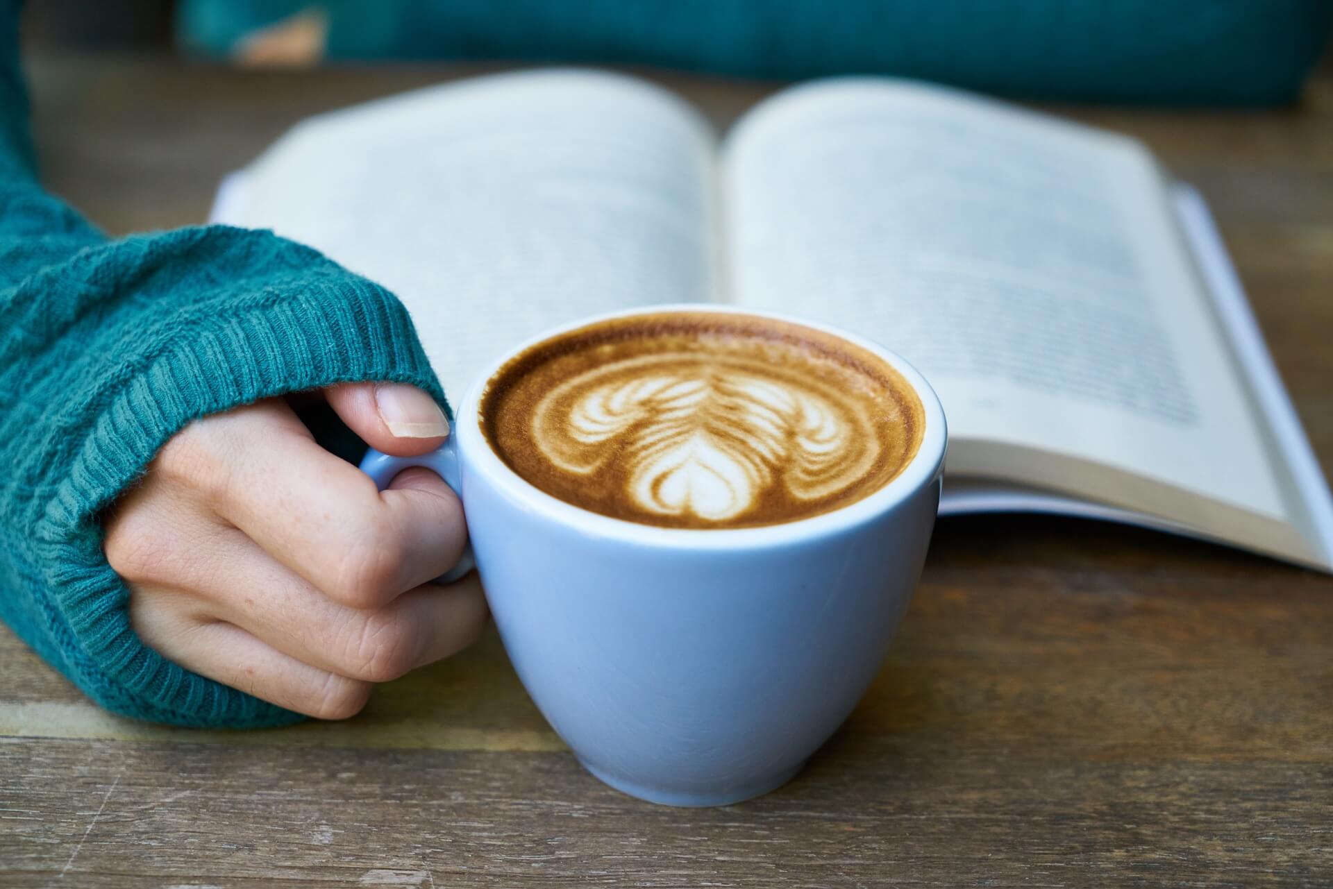 A hand holding a cup of coffee with a book on a table in the background.